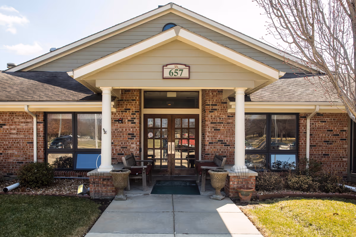 Front entrance of a brick building with a peaked roof and two white columns supporting a small porch. There are two wooden benches with red cushions on either side of the entrance door. The building number 657 is displayed above the door. There are windows on both sides of the entrance and some landscaping with bushes and grass in front.