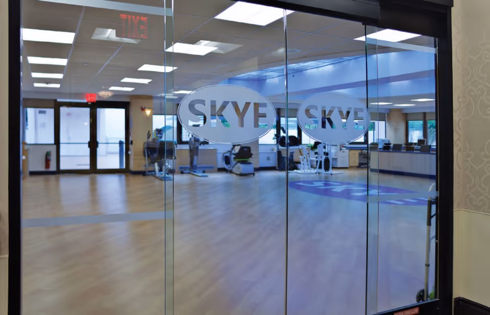 View through glass doors labeled 'SKYE' into a spacious fitness room with exercise equipment, wooden flooring, and bright overhead lighting.