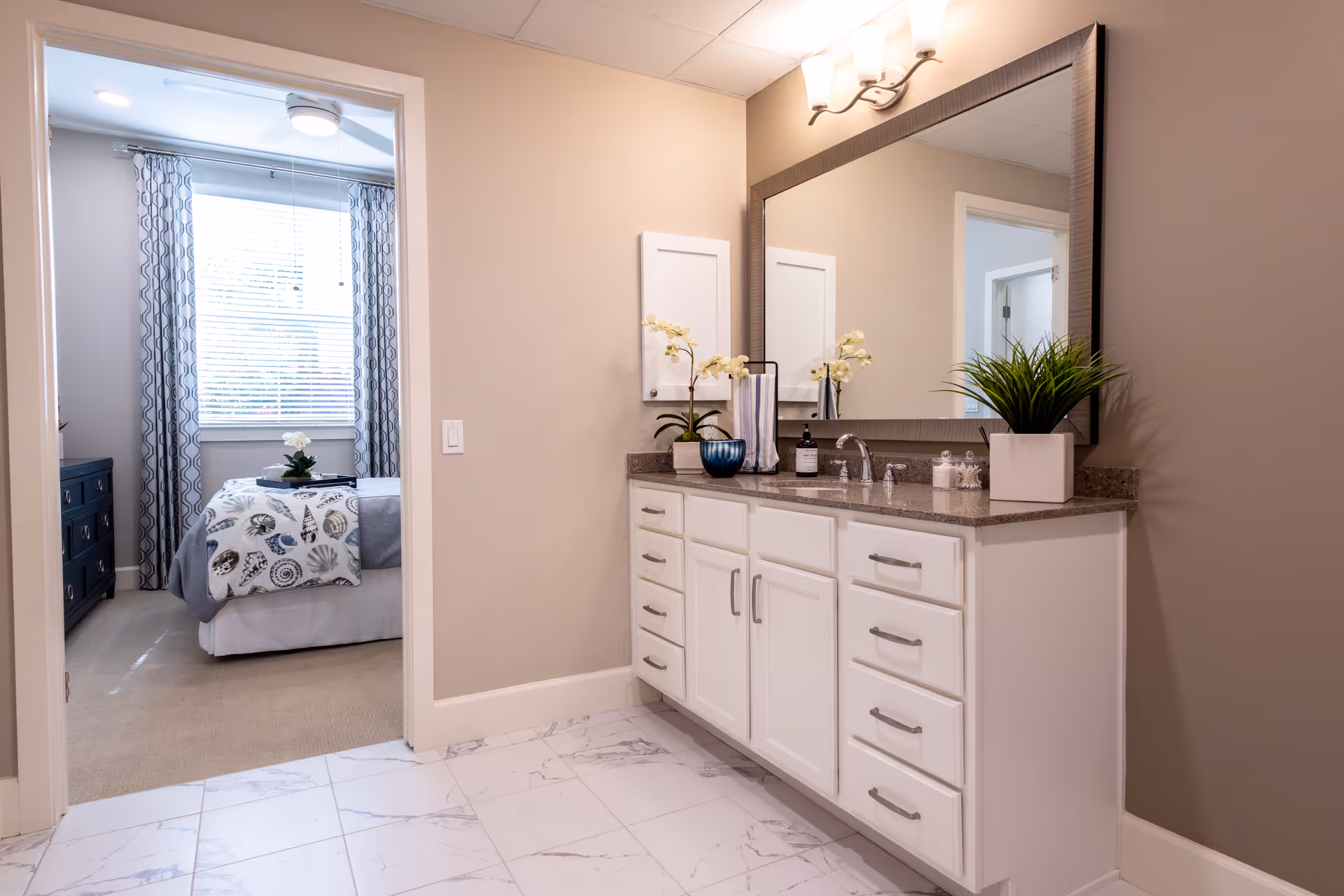 Clean bathroom with white cabinets, marble floor, large mirror, and a doorway to a bedroom.