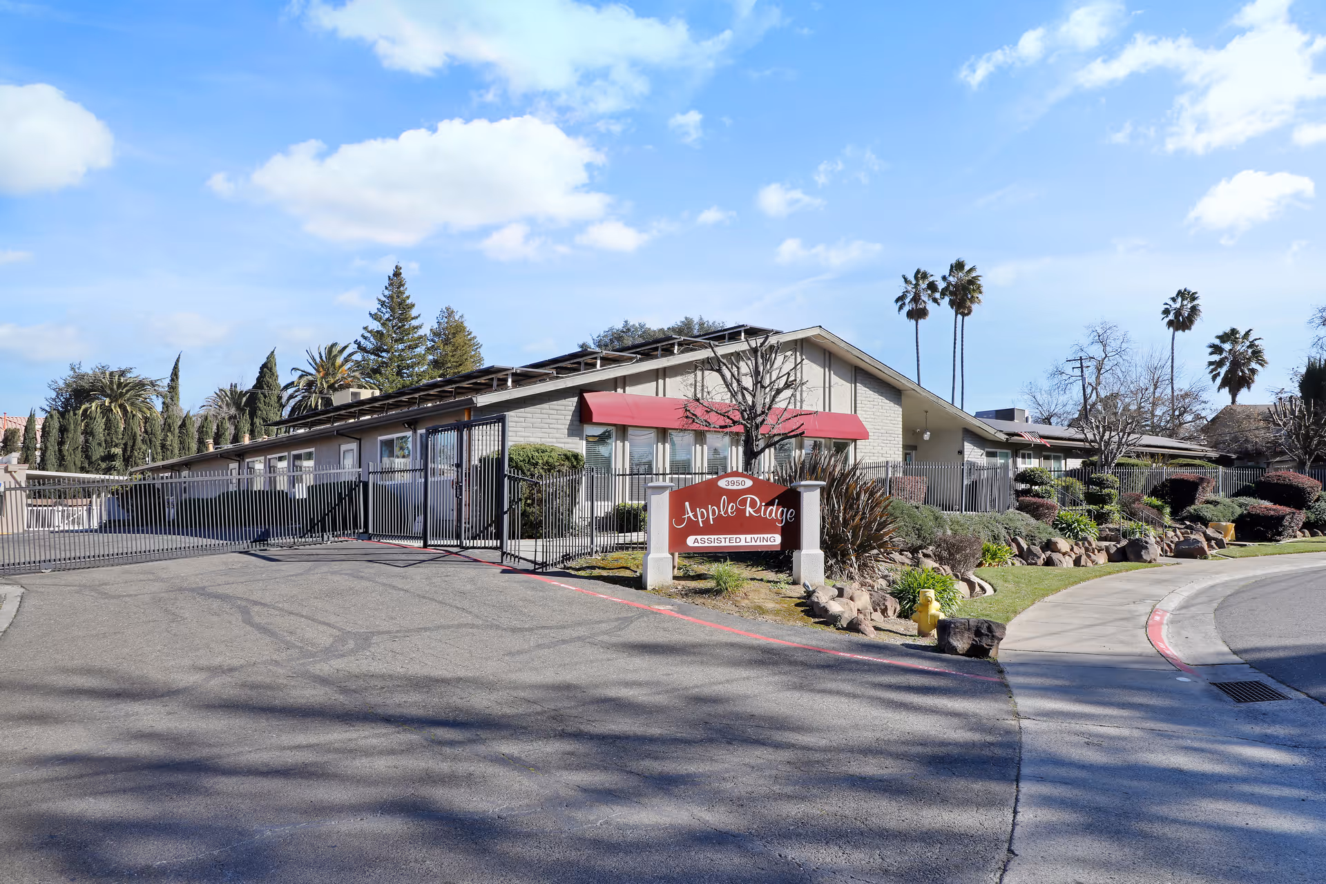 Exterior view of Apple Ridge Assisted Living facility showing a single-story building with a gated entrance, a red awning above the windows, landscaped bushes and trees, a curved sidewalk, and a clear blue sky with some clouds.