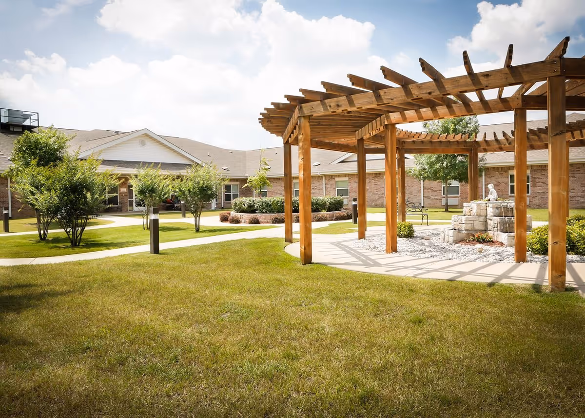Outdoor courtyard of a senior home with a wooden pergola, walking paths, and a stone fountain.