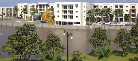 View of a modern multi-story senior living facility named Amavida Living - Water's Edge, situated along a waterfront with palm trees and other greenery in the foreground, and a calm body of water separating the building from the landscaped area.