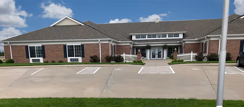Front exterior view of a single-story brick building with a gray shingled roof, white trim, and black shutters. The entrance features double glass doors with a small covered porch supported by columns and decorated with hanging plants. There is a paved parking area with marked spaces and a green lawn in the foreground under a partly cloudy blue sky.
