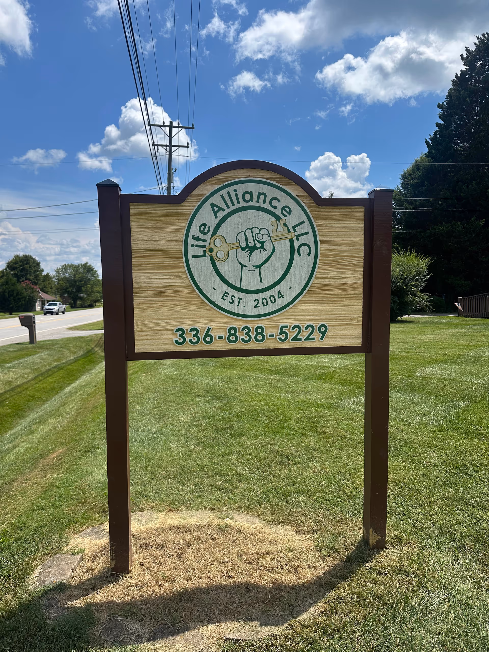 Outdoor Life Alliance LLC sign on grassy verge beside road, featuring emblem and contact number.