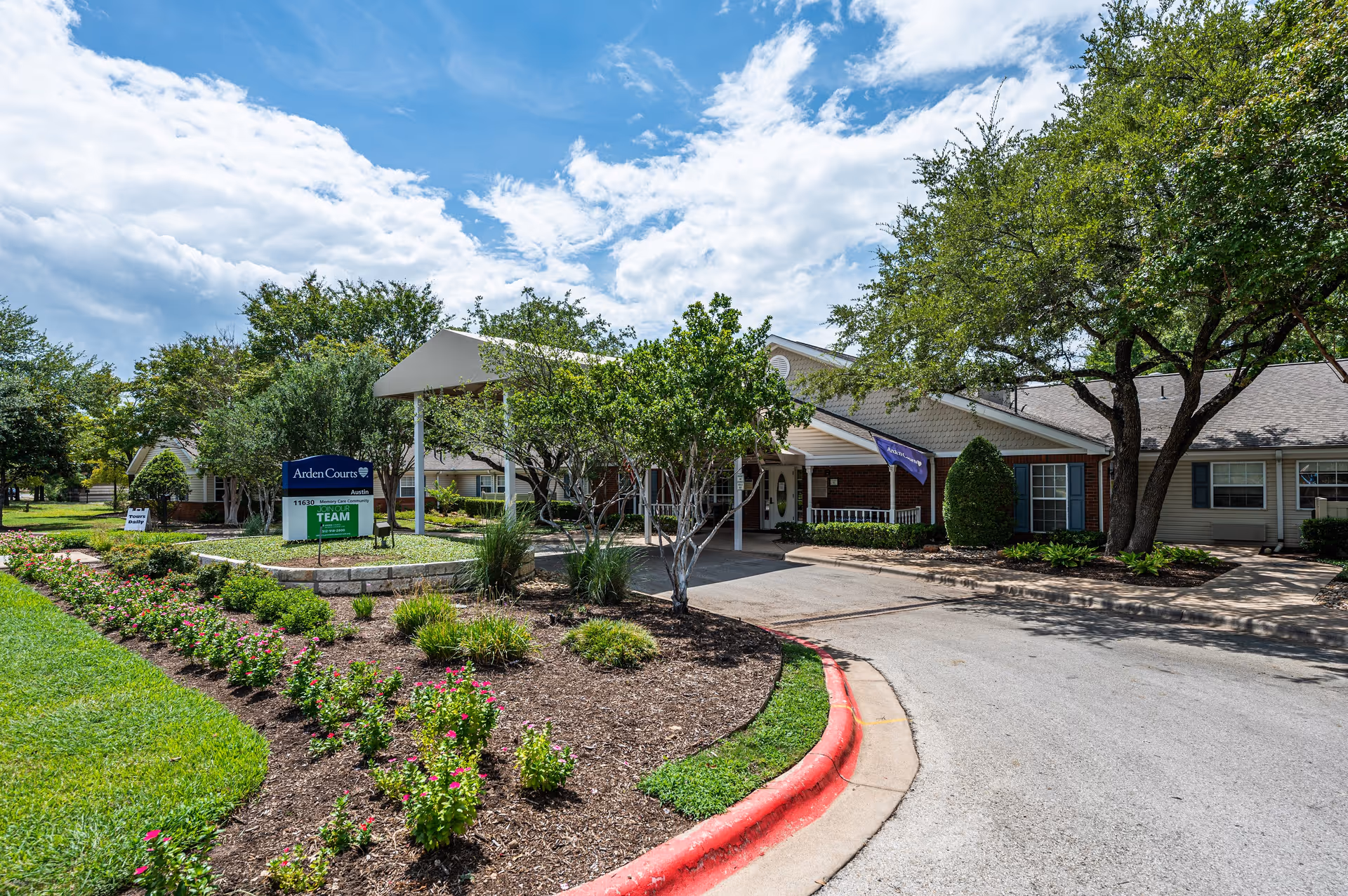 Exterior view of Arden Courts - ProMedica Memory Care Community in Austin, showing the entrance with a covered driveway, landscaped garden with flowers and trees, and a clear blue sky with some clouds.