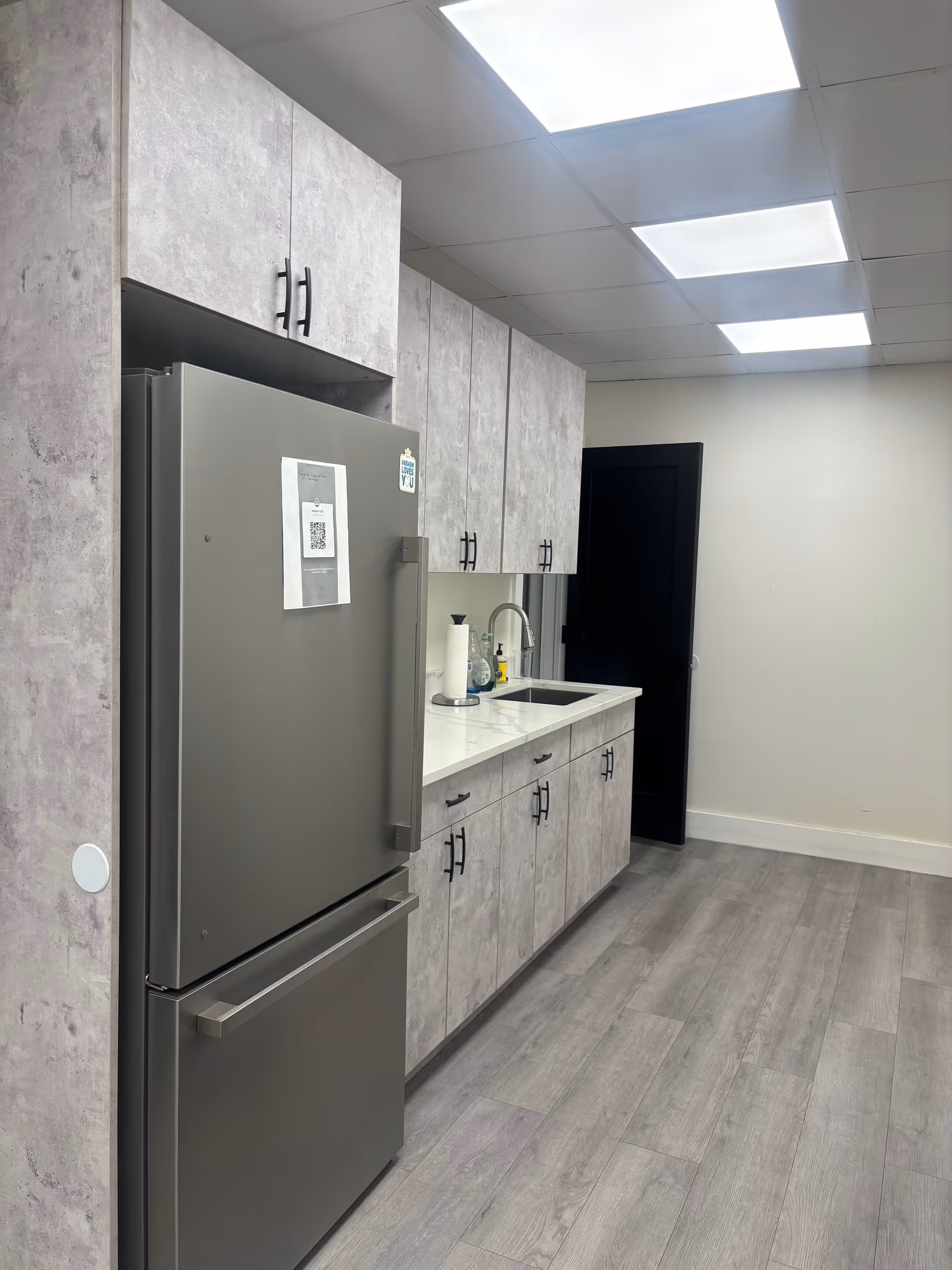 Modern kitchenette with gray cabinets, stainless refrigerator, sink, and bright ceiling lights.