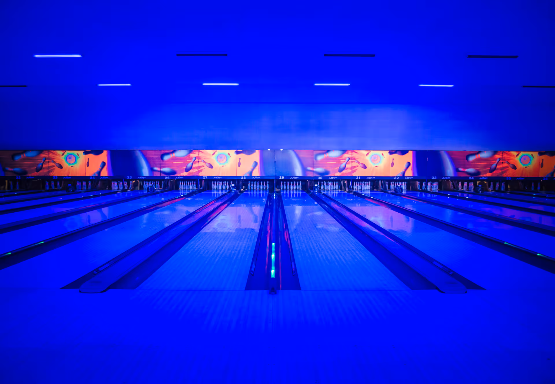Blue-lit indoor bowling alley with numerous lanes and colorful wall art.