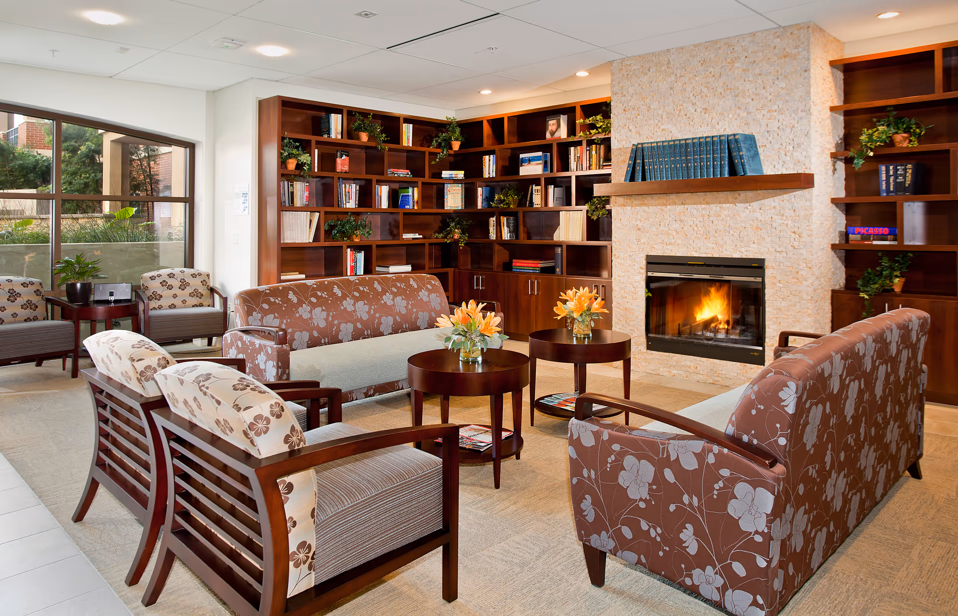 A cozy living room area with floral patterned sofas and armchairs arranged around two round wooden coffee tables with flower vases. A stone fireplace with a fire burning is centered on the wall, flanked by built-in wooden bookshelves filled with books and plants. Large windows on the left side let in natural light and show greenery outside.