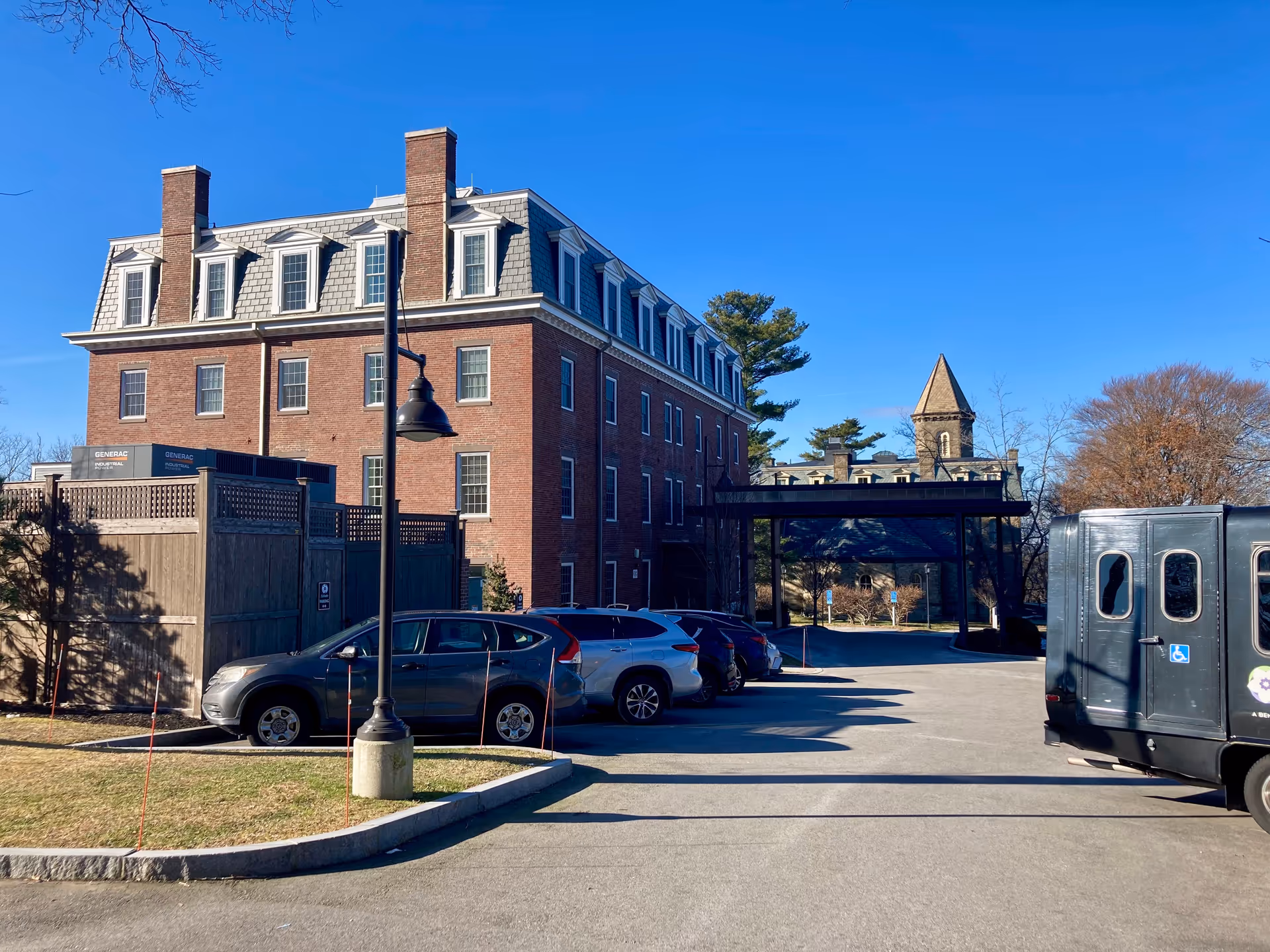 Exterior view of a multi-story brick building with a mansard roof and multiple windows, adjacent to a parking lot with several parked cars and a black vehicle with handicap signs. The sky is clear and blue, and there are trees in the background.