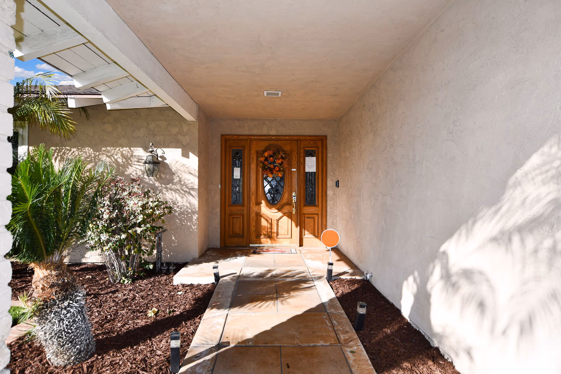 Wooden front door under a covered entrance, with landscaping and a paved walkway.