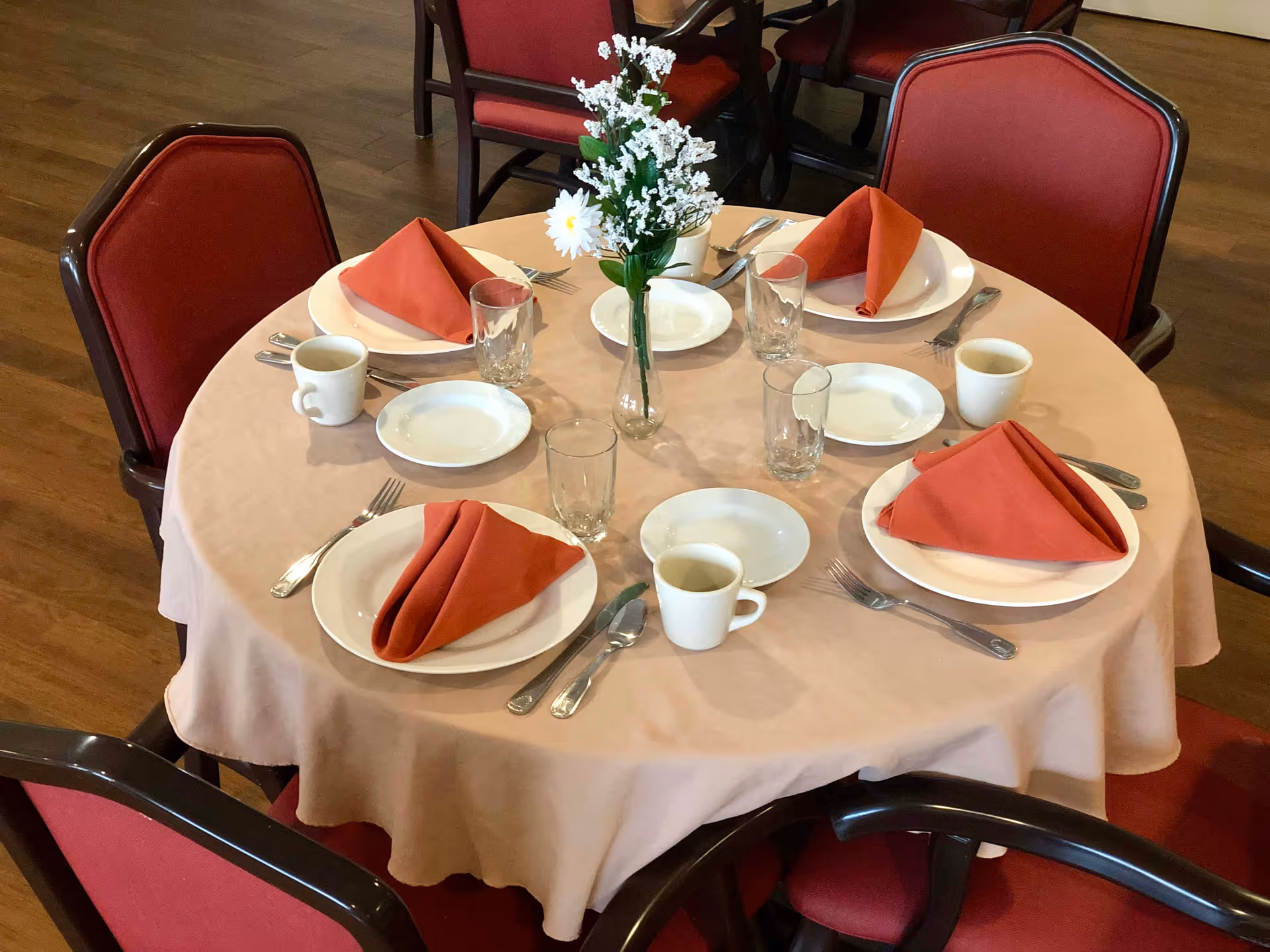 Round dining table in a communal dining room set for five with plates, glasses, coffee cups, folded red napkins and a small floral centerpiece.