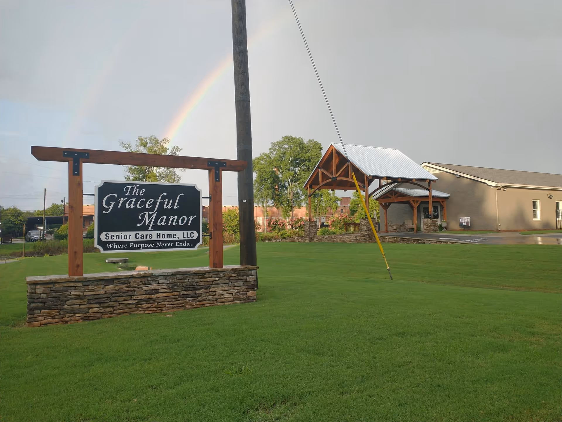 Outdoor view of The Graceful Manor Senior Care Home sign on a stone base with a green lawn in front. In the background, there is a building with a covered entrance and a double rainbow in the sky.