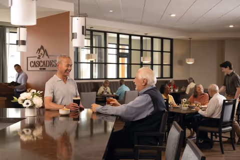 Two elderly men sitting at a bar counter in a senior living community dining area, smiling and holding drinks. Other seniors are seated at tables in the background, engaged in conversation and dining. The room has modern lighting and large windows with black frames.