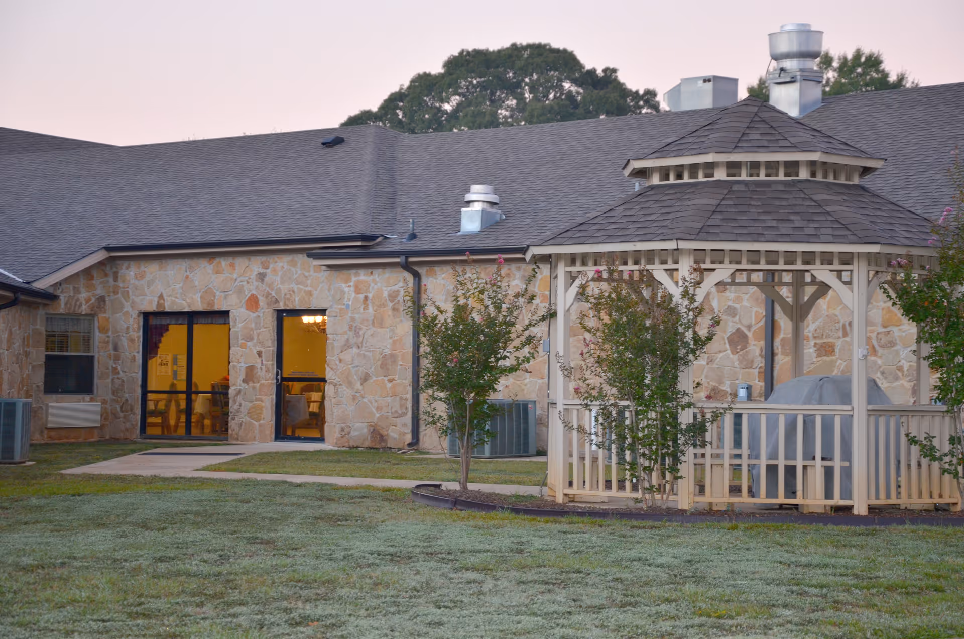 A wooden gazebo sits on a grassy courtyard in front of a stone-faced nursing facility building with lit windows.