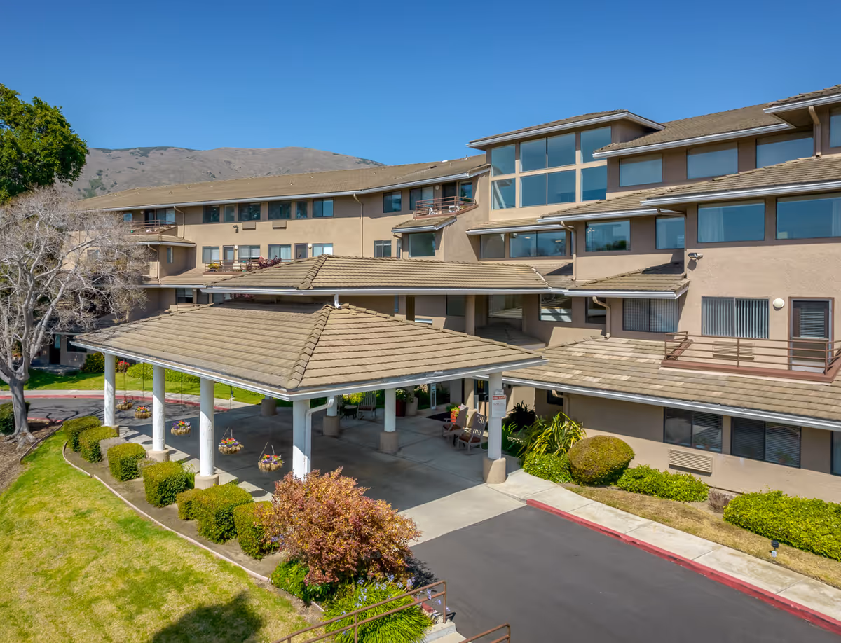 A tan multi-story senior living facility with a covered entrance and landscaped grounds.
