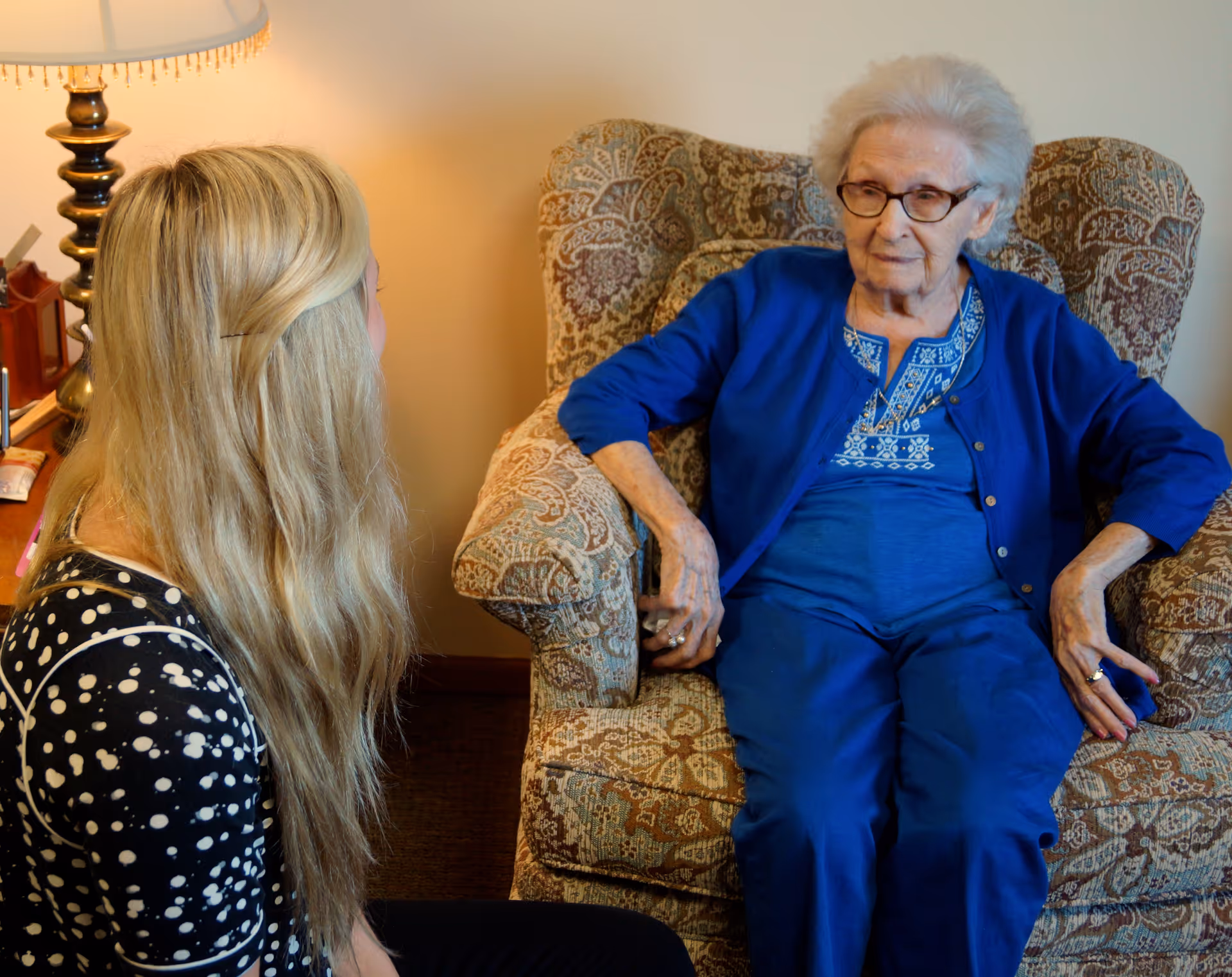 An elderly woman wearing glasses and a blue outfit sits in a patterned armchair while a younger woman with long blonde hair, wearing a black and white polka dot top, sits nearby facing her. A lamp with a white shade is visible in the background.
