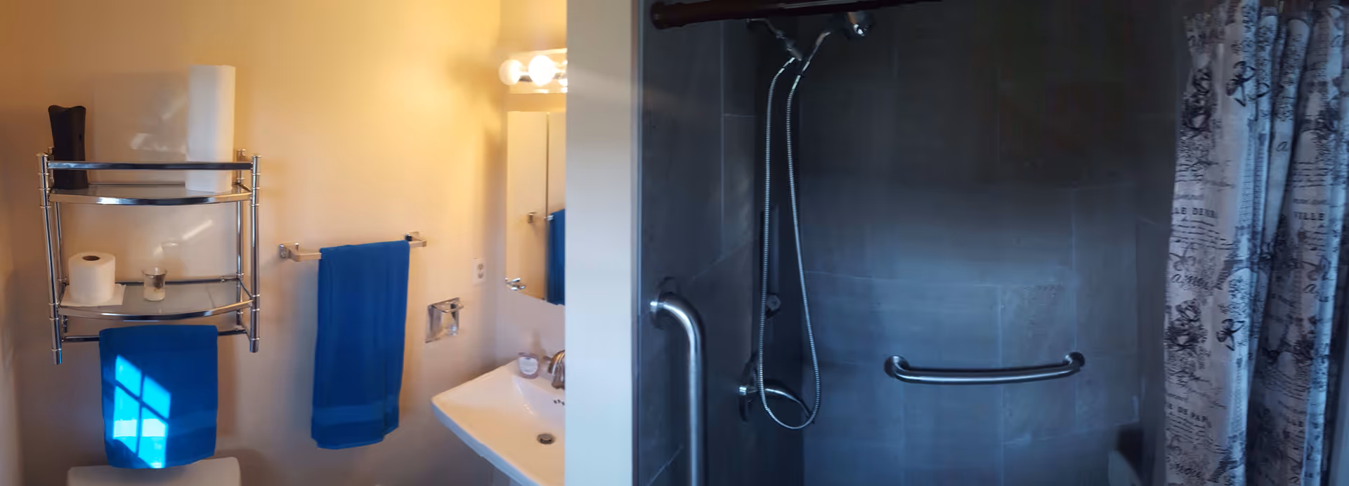A bathroom with a white sink, a mirror with lights above it, a metal shelf holding toilet paper and a candle, blue towels hanging on towel racks, and a shower area with dark tiles, a handheld showerhead, grab bars, and a patterned shower curtain.
