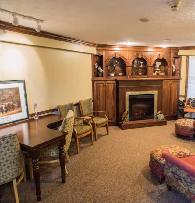 A cozy living room area with a wooden electric fireplace surrounded by a built-in wooden cabinet displaying decorative items. There are several upholstered chairs and a patterned ottoman arranged around the room, with a wooden table and framed artwork on the left side. The room has beige walls and carpeted flooring.
