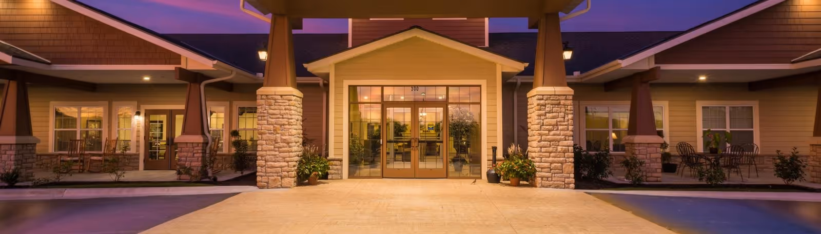 Front exterior view of Oak Pointe of Carthage facility at dusk with warm lighting, stone pillars, glass entrance doors, and outdoor seating areas on both sides.