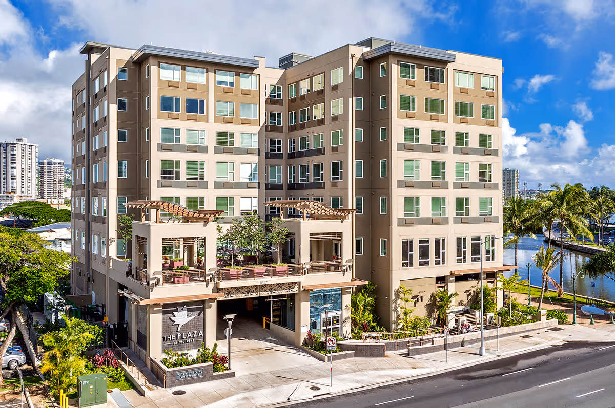 Exterior front view of a multi-story beige senior living building labeled "The Plaza" with balconies, landscaping, and a waterfront nearby.