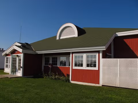 Red single-story building with white trim, green roof, arched front dormer, and lawn.