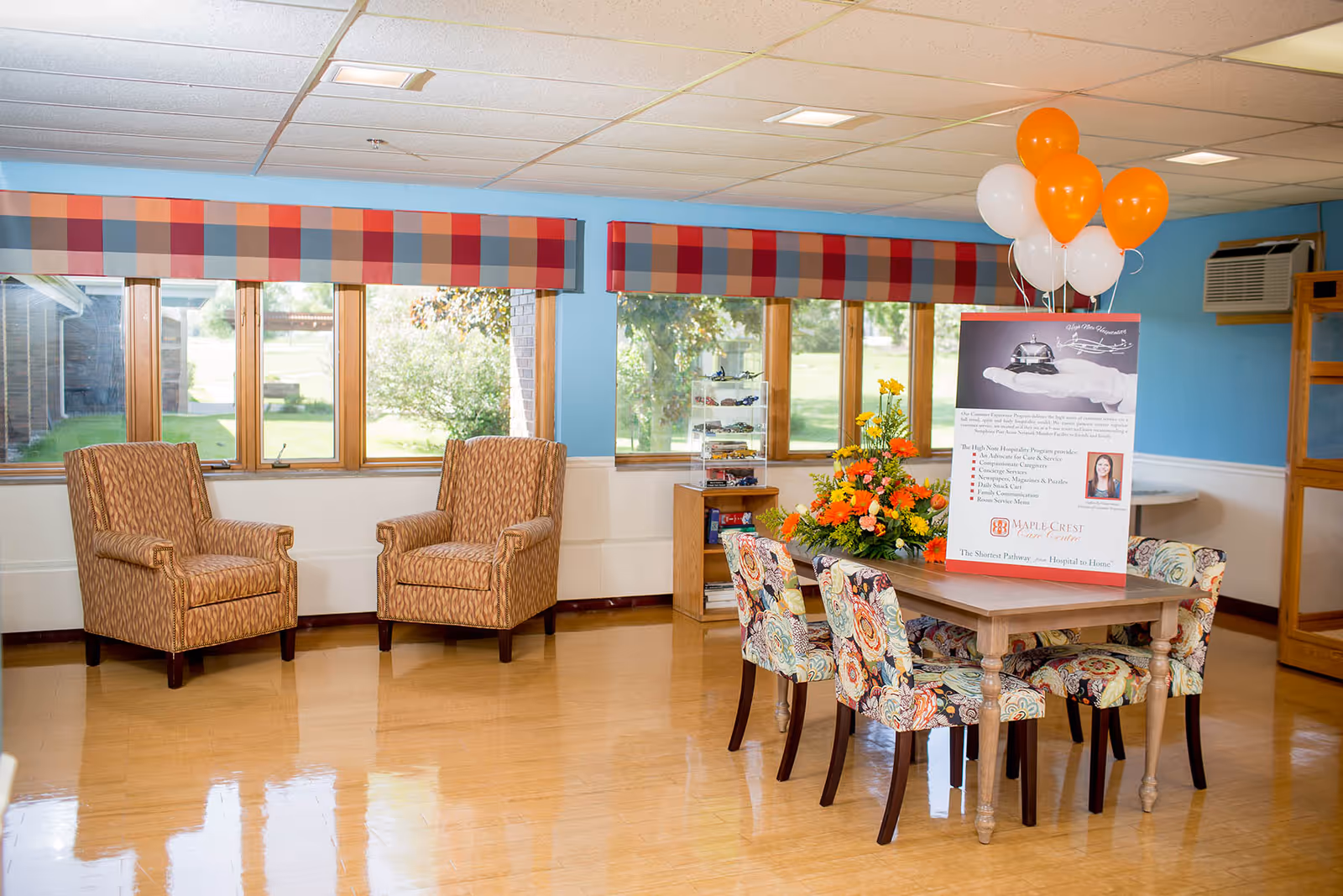 Bright facility lounge with two armchairs, a decorated table, flowers, balloons, and a promotional display.