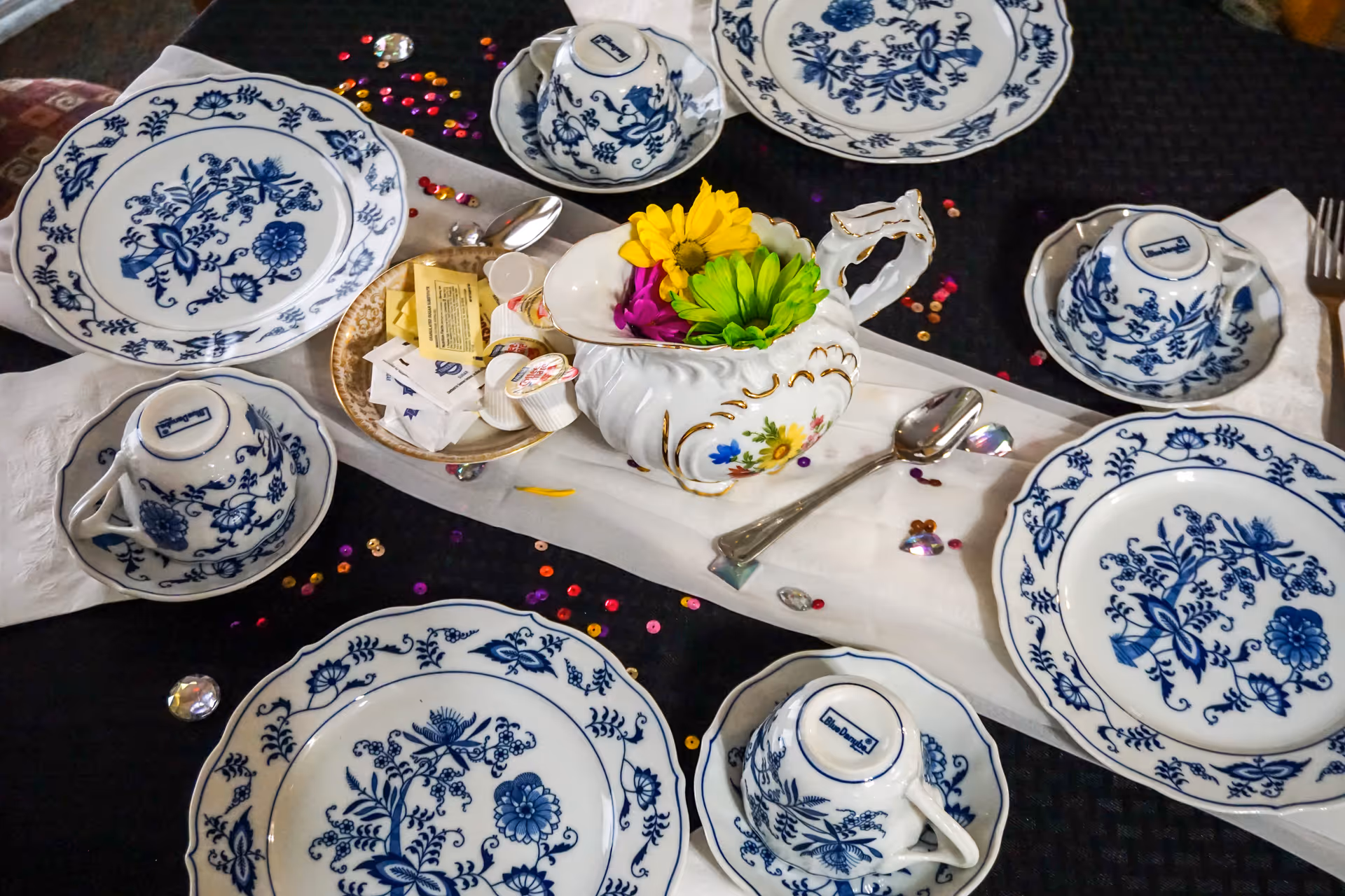A dining table set with blue and white floral patterned plates, teacups, and saucers. The table has a black tablecloth with a white runner, decorated with colorful sequins and a small white vase holding yellow, green, and purple flowers. There are also small containers of cream and sugar packets on the table.