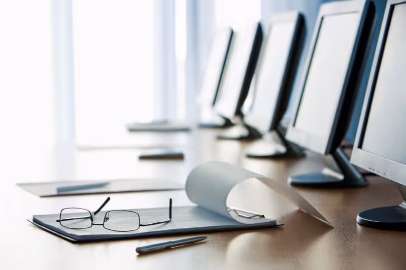 Row of computer monitors on a desk with glasses and papers.