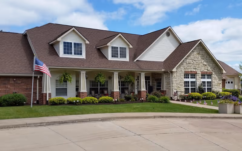 Exterior view of a single-story senior living facility building with a combination of brick and stone facade, a brown shingled roof, and white trim. The building features a covered porch with columns, hanging plants, and rocking chairs. An American flag is displayed on a flagpole in the front lawn, which is landscaped with bushes and flowers under a partly cloudy sky.