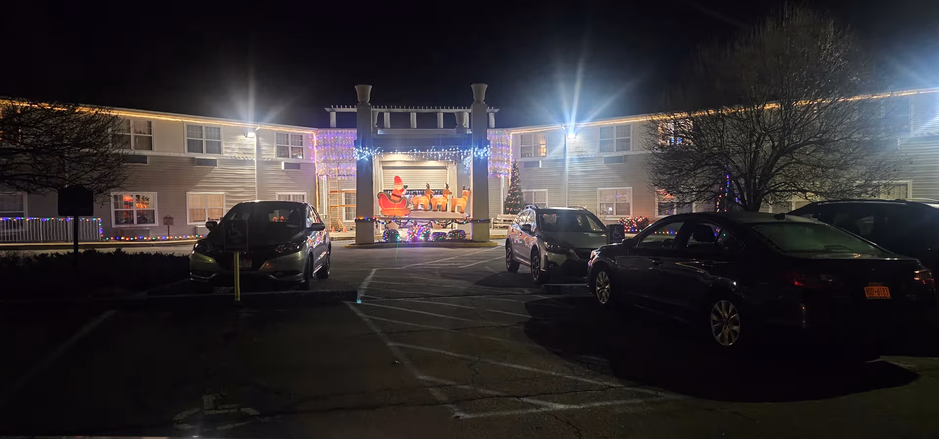 Night view of The Plaza at Clover Lake exterior with holiday lights and parked cars.