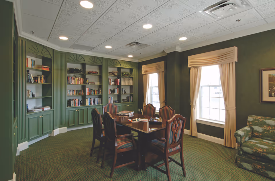 A cozy interior room with green walls and carpet, featuring built-in bookshelves filled with books and decorative items. In the center, there is a wooden table surrounded by six upholstered chairs with striped fabric. Two windows with beige curtains allow natural light into the room. A floral-patterned armchair is partially visible on the right side.