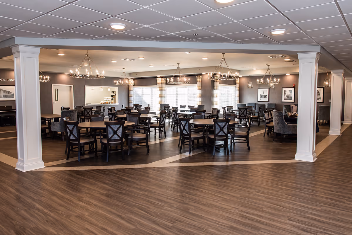 Spacious dining hall with multiple tables, chairs, and chandeliers in a senior living facility.