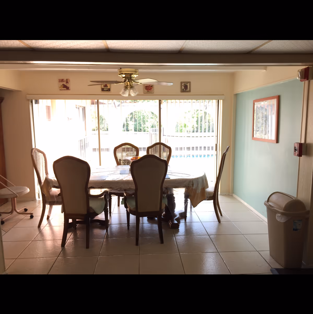 Dining room with a table covered by a tablecloth and six chairs under a ceiling fan, facing sliding glass doors that open to a pool area.