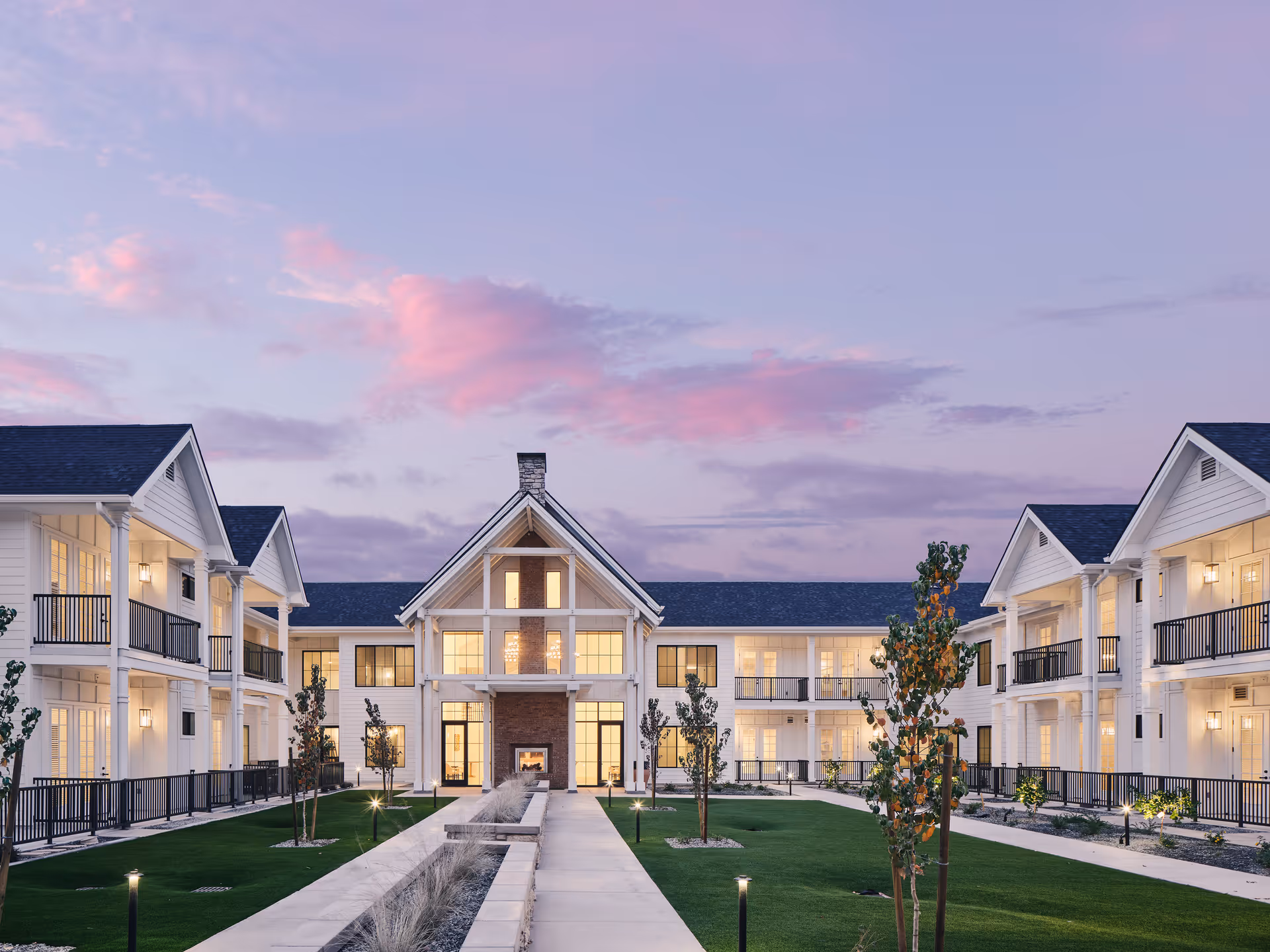 Courtyard view of a two-story white senior living building with balconies, a central entrance, and landscaped lawn under a pink-purple sky.