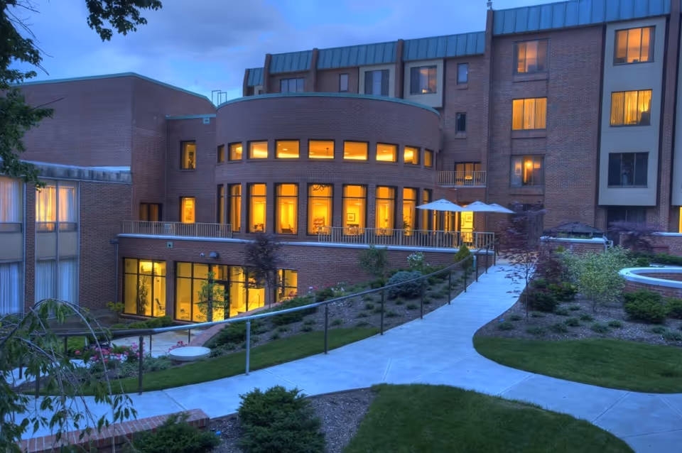 Exterior view of a multi-story brick retirement community building at dusk with warm interior lights, a curved walkway, and landscaped courtyard.