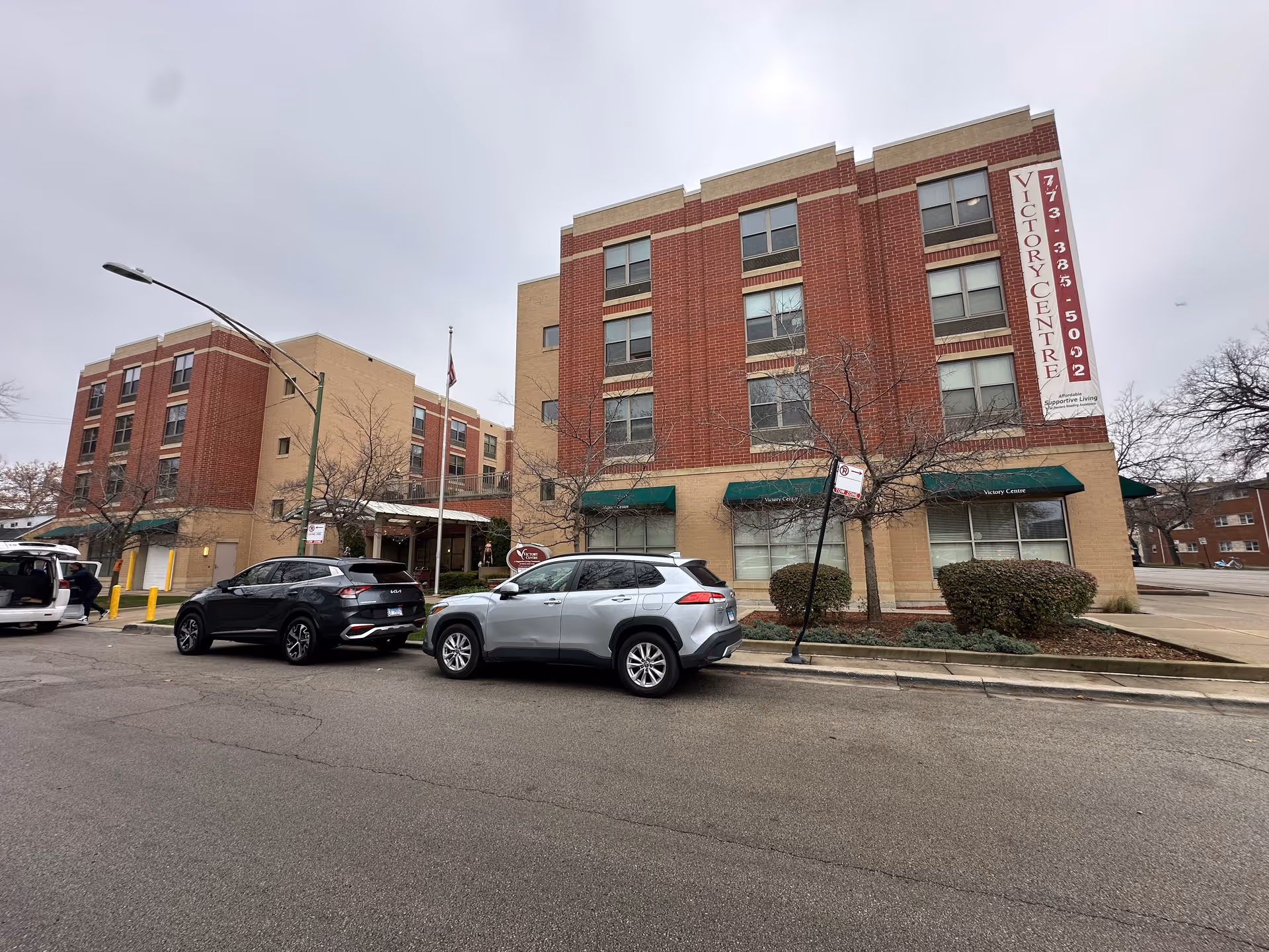 Exterior view of a multi-story brick-and-tan senior living building with green awnings and cars parked along the street.