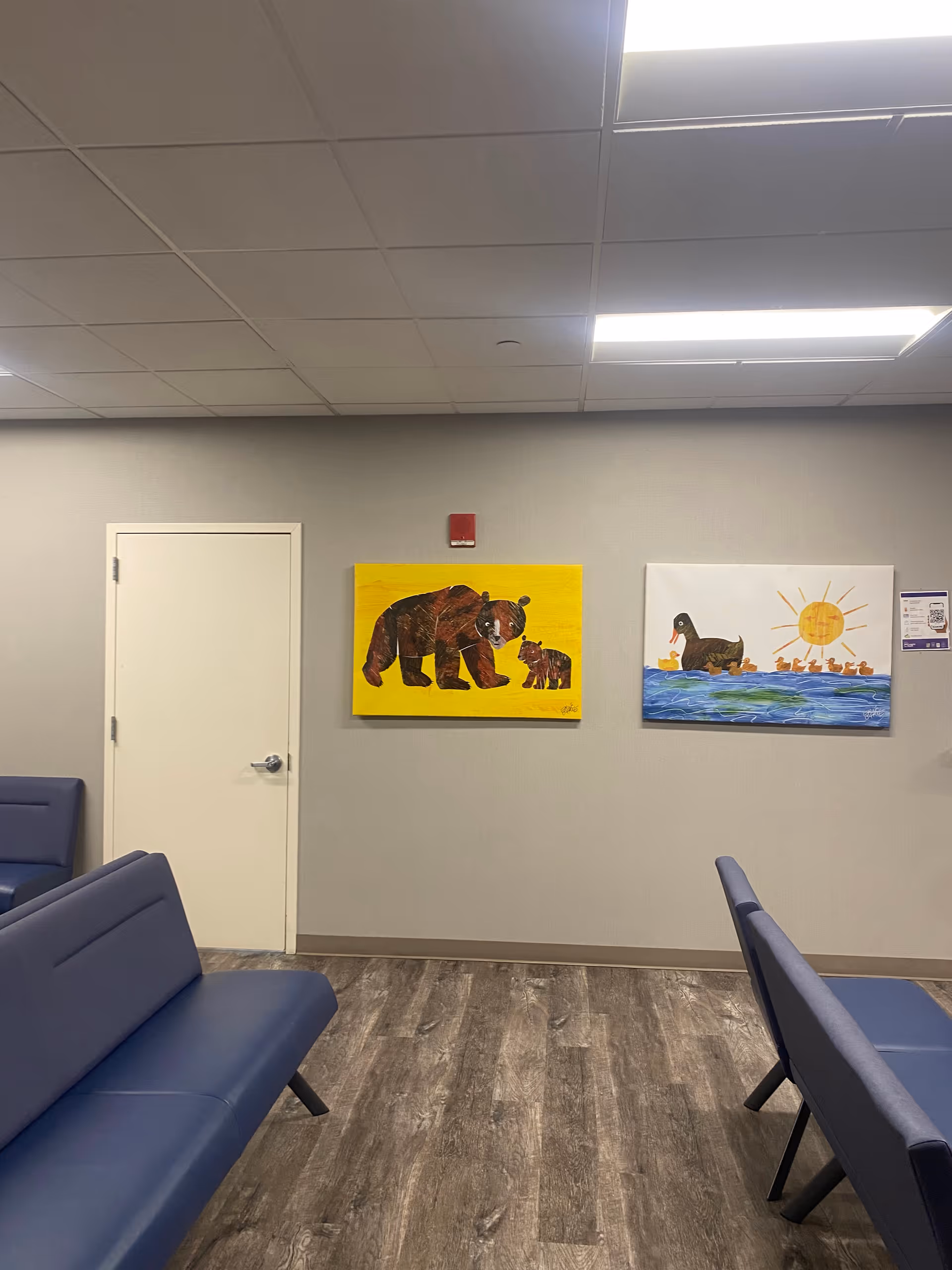 Indoor waiting area with blue benches, two wall paintings, a white door, and wood-look flooring.