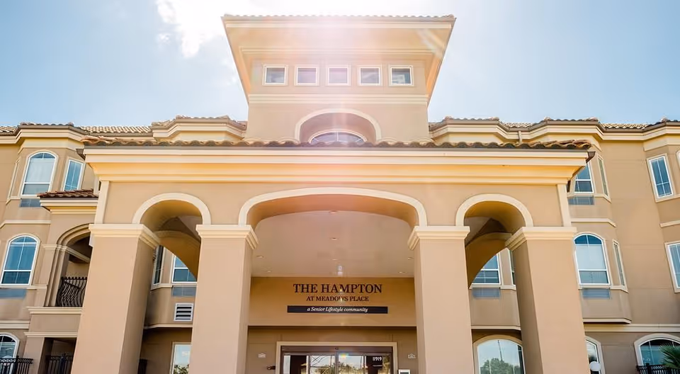 Sunlit front entrance of The Hampton at Meadows Place senior living building with an arched portico and multiple windows.