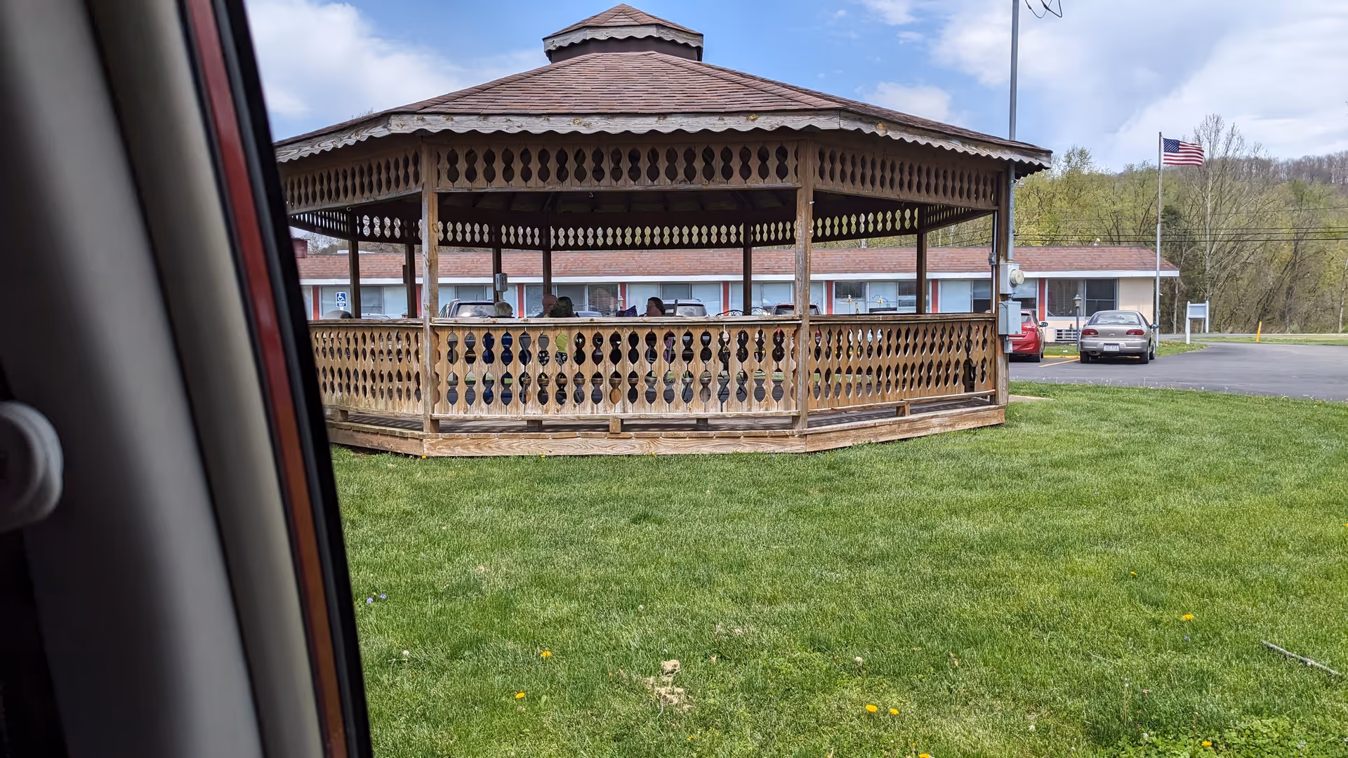 Wooden gazebo on a grassy lawn with a building and parked cars nearby.