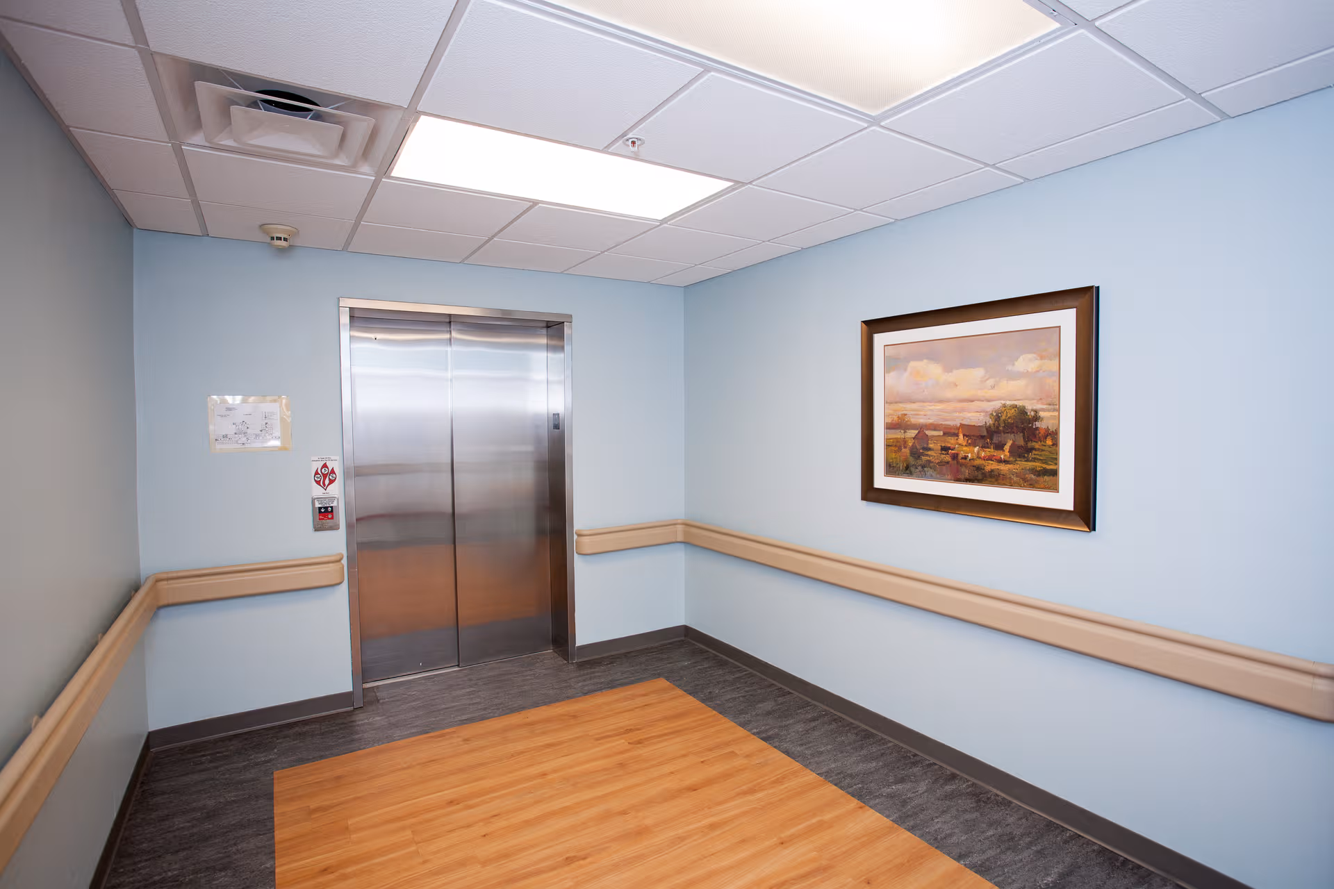 Elevator door in a light-blue hallway with framed artwork and wall railings.