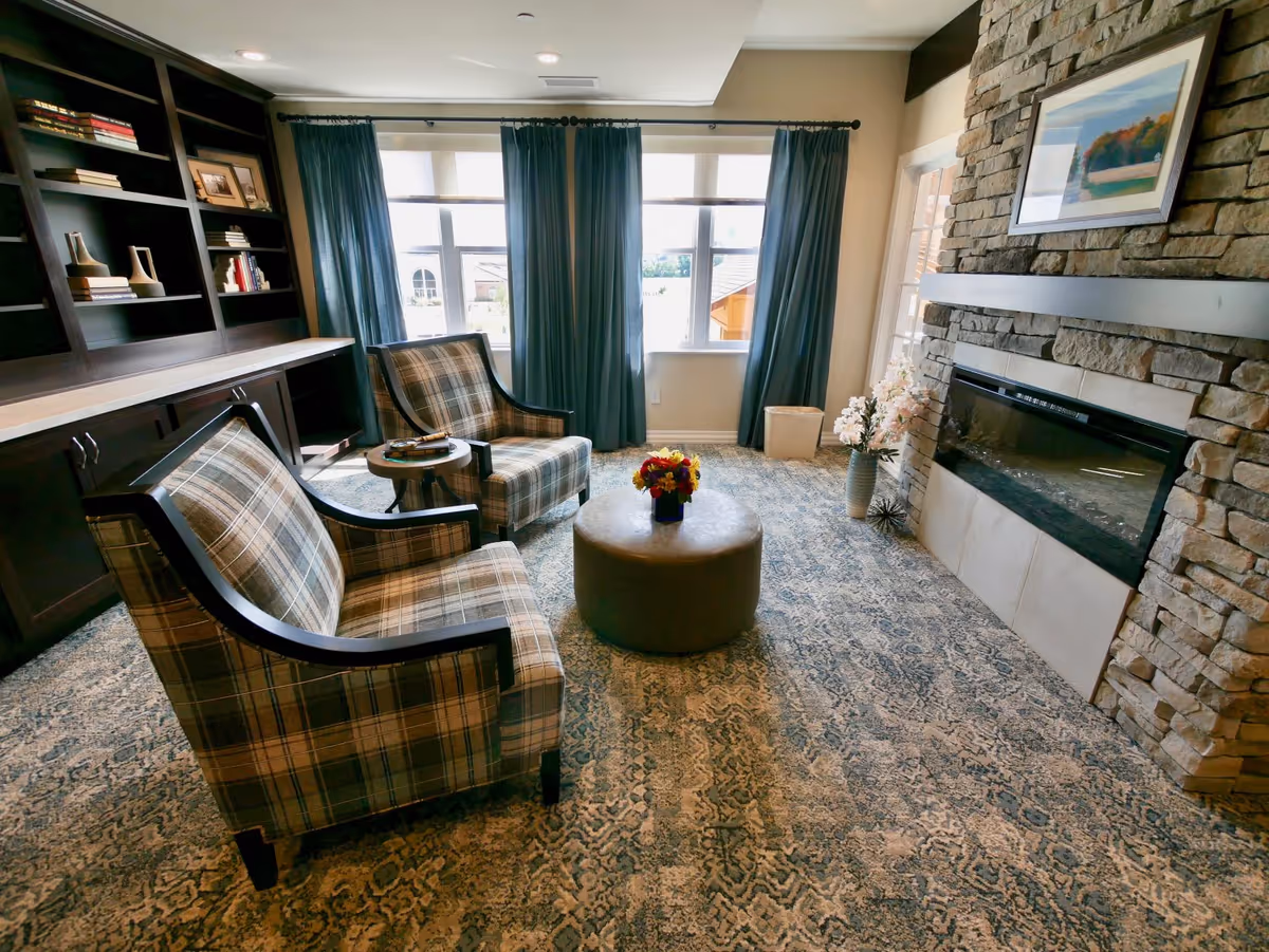 Cozy seating area with two plaid armchairs around a round ottoman topped with flowers, built-in bookshelves, large windows with curtains, and a stone fireplace.