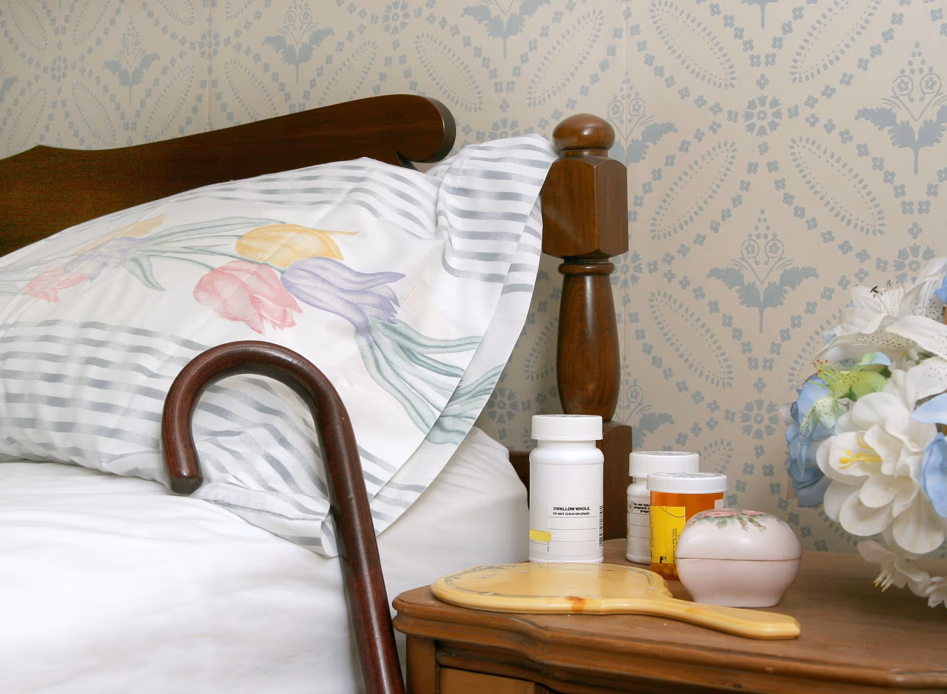 Bed with floral pillow, pills on nightstand, and floral wallpaper in a bedroom.
