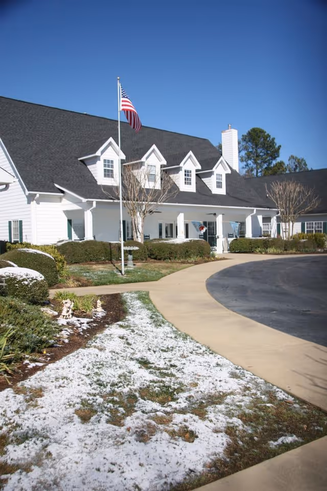 Exterior view of a white senior living facility building with a black roof, an American flag on a flagpole, trimmed bushes, and a curved driveway. There is a light dusting of snow on the grass and garden beds under a clear blue sky.