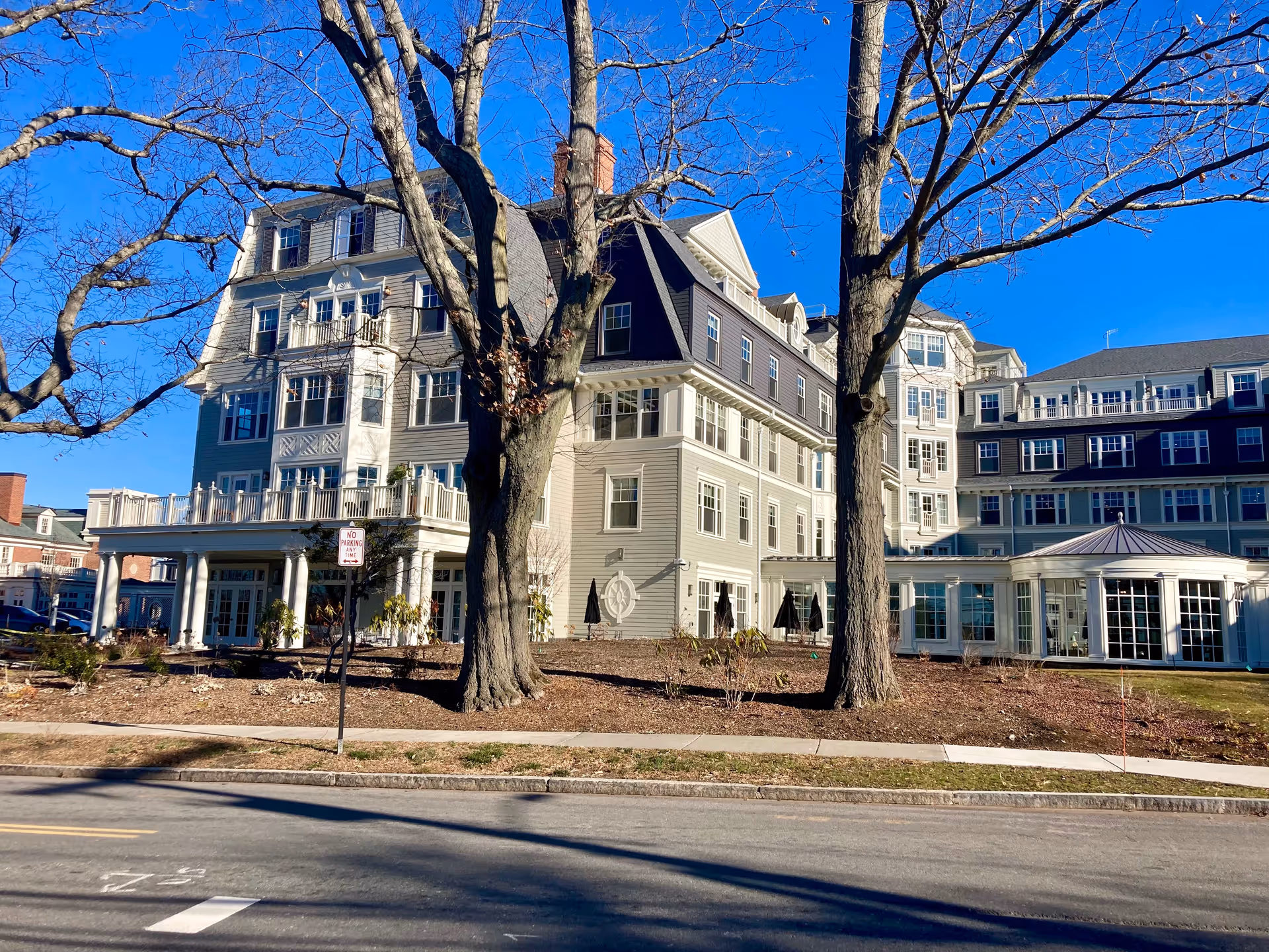 Multi-story colonial-style senior living building with porches and large bare trees in front on a sunny day.
