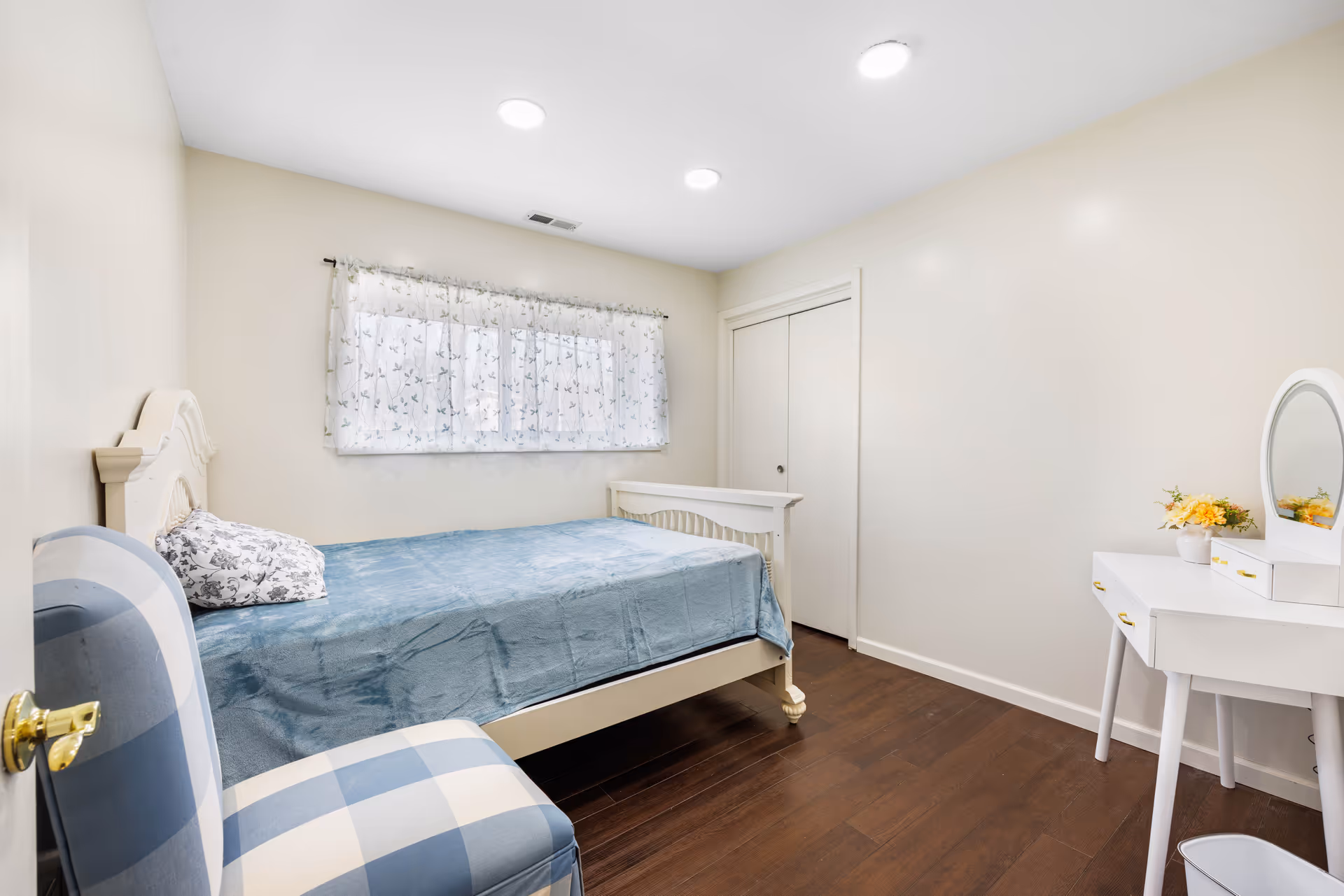 A small, neatly furnished bedroom with a bed, vanity, dresser, and window curtains.