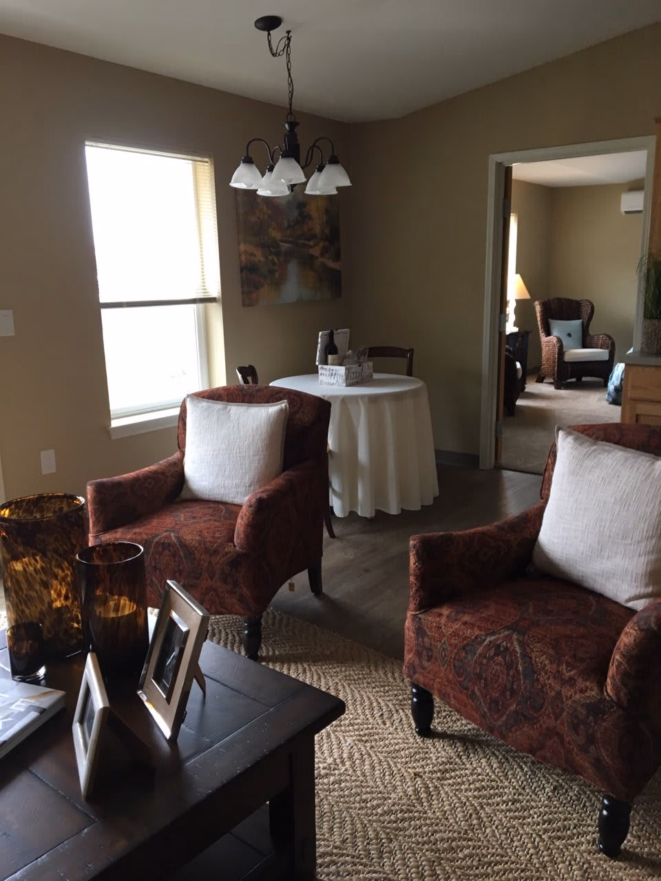 Interior view of a senior living apartment showing a cozy living room area with two patterned armchairs with white cushions, a dark wooden coffee table with decorative vases and photo frames, a round dining table covered with a white tablecloth, and a window letting in natural light. In the background, there is an open doorway leading to another room with a wicker armchair and a lamp.