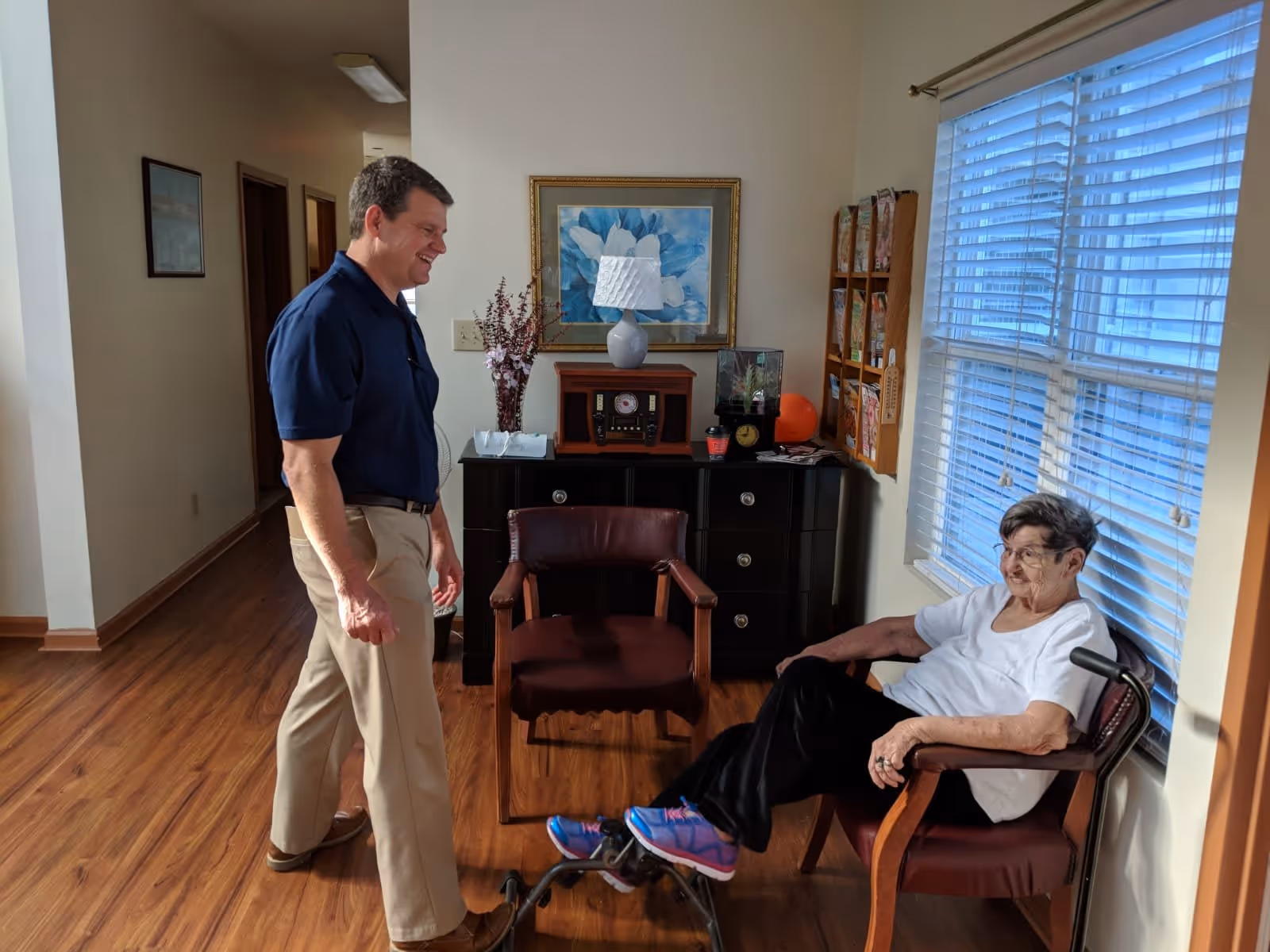 An elderly woman sitting in a chair next to a window with blinds, smiling and interacting with a man standing nearby in a room with wooden flooring and a black cabinet with decorative items.