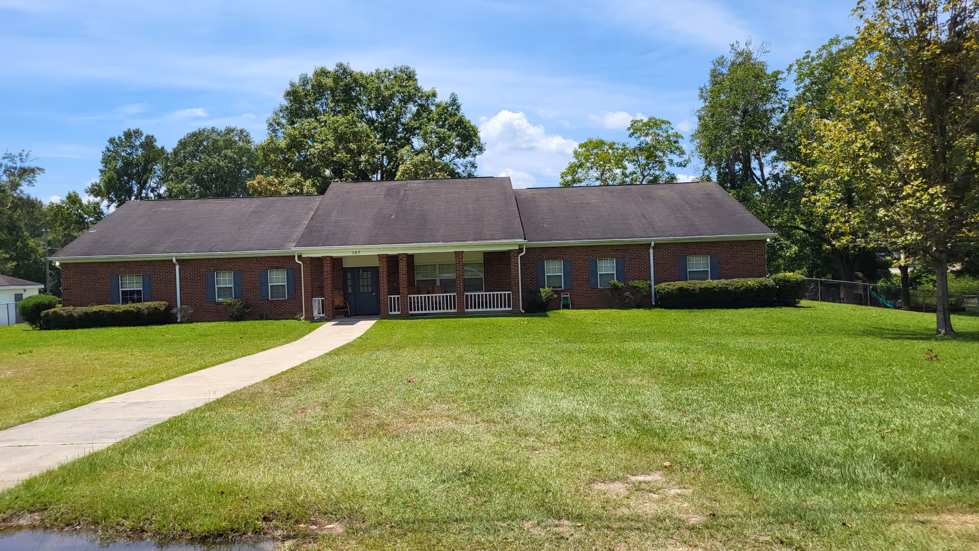 A brick, single-story building with a front porch, grassy lawn, and surrounding trees.