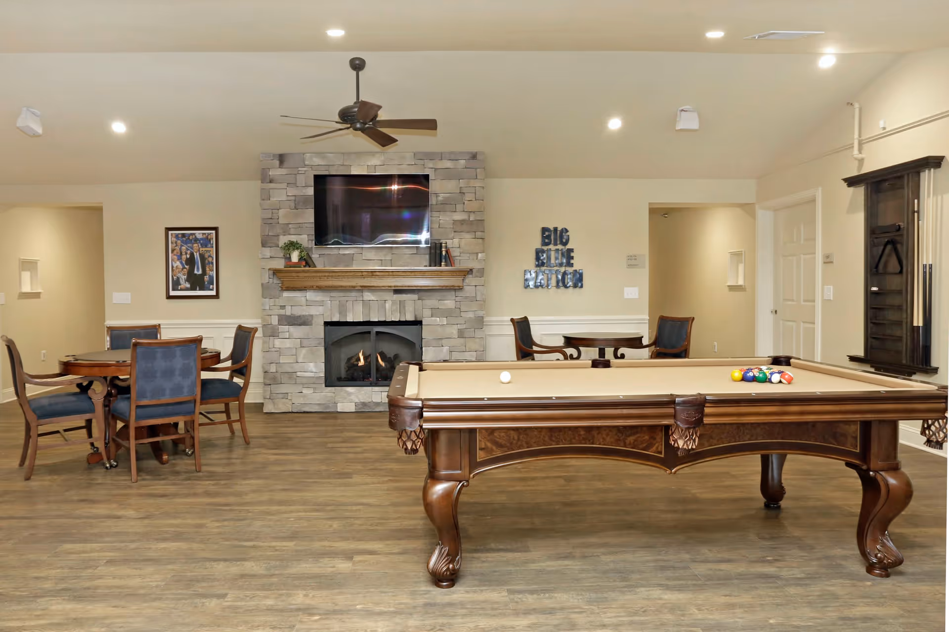 A recreational room with a pool table in the foreground, a stone fireplace with a mounted TV above it, and two round tables with chairs around them. The walls are light-colored, and there is a ceiling fan above the fireplace. On the right wall, there is a sign that reads 'BIG BLUE NATION'.