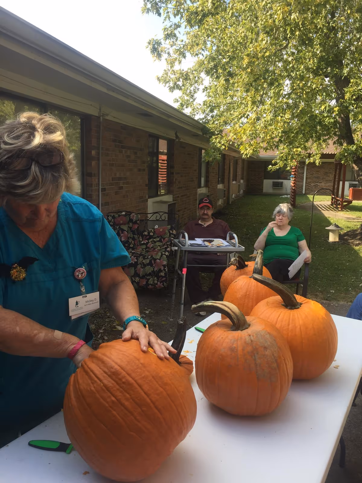 A woman in a teal uniform is carving a large pumpkin on a white table outdoors. Three other large pumpkins are on the table. In the background, two elderly people are seated near a brick building with windows, under a tree with green leaves.